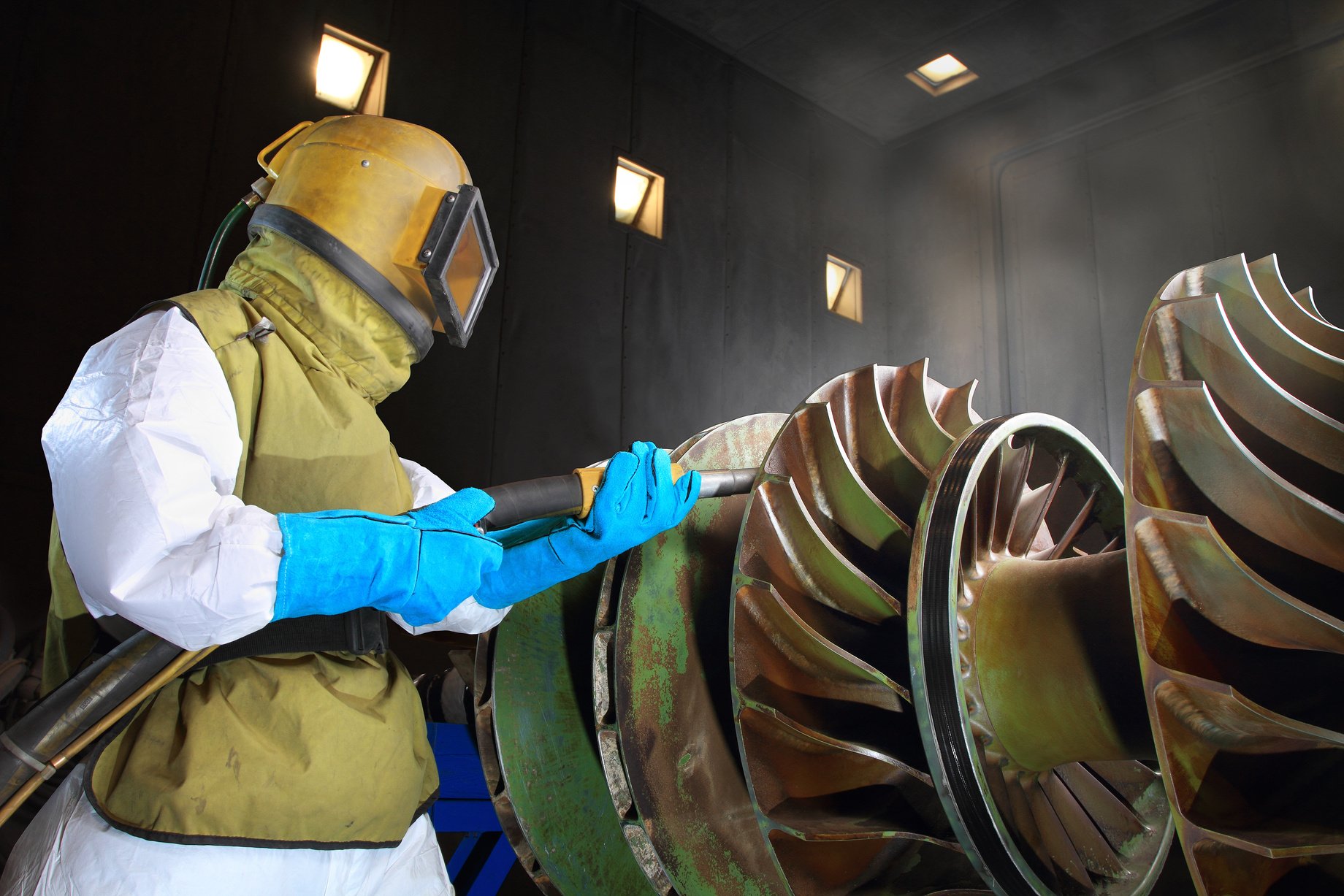 Man in protective gear sandblasting a giant metal wheel